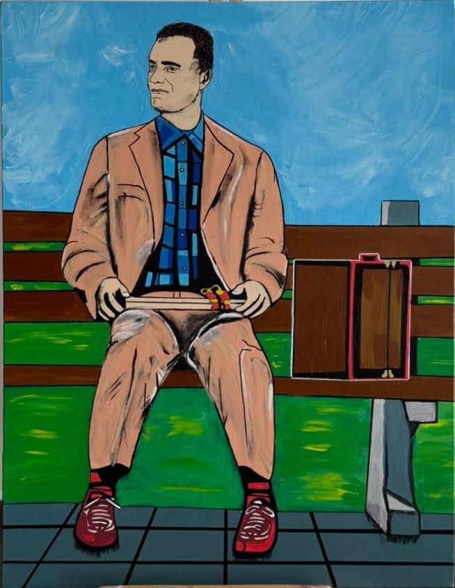 Homme en costume beige assis sur un banc, une boîte de chocolats sur les genoux et des chaussures rouges aux pieds.